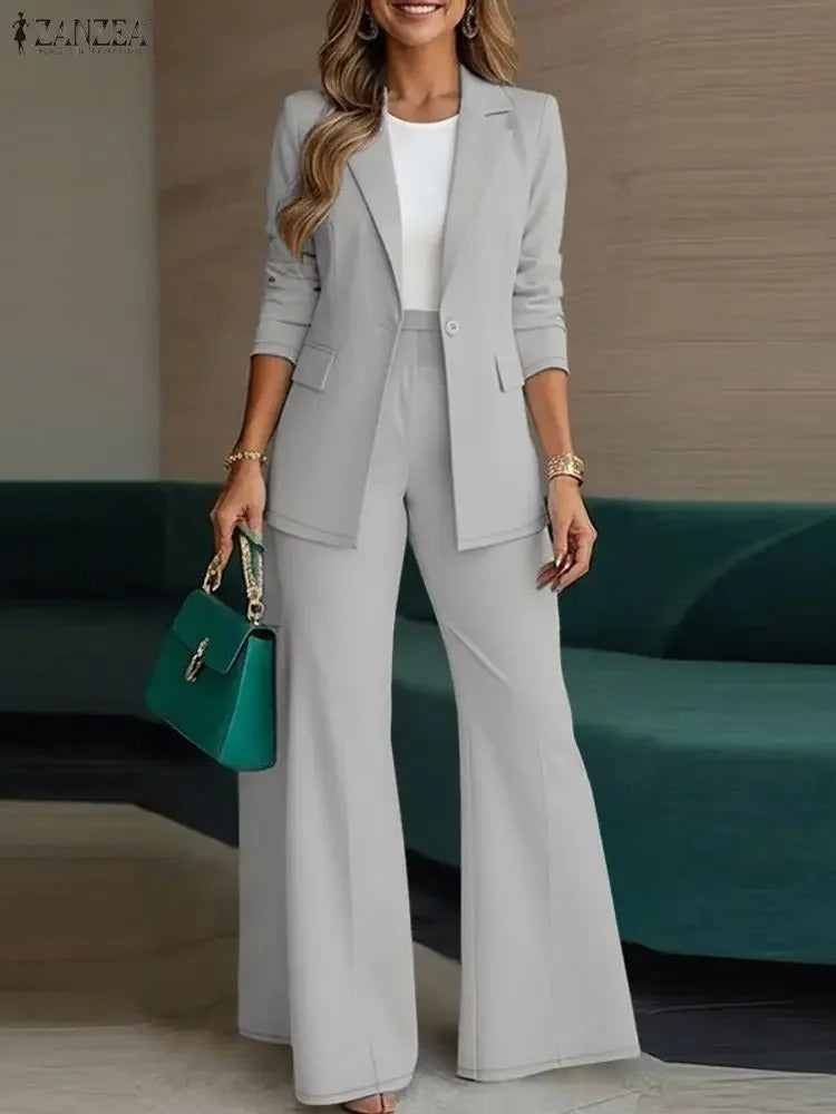 ZANZEA fashion photography featuring a smiling woman in a stylish light gray pantsuit with wide-leg trousers and a white top, accessorized with an emerald green handbag and gold bracelets, posing indoors by a dark teal sofa